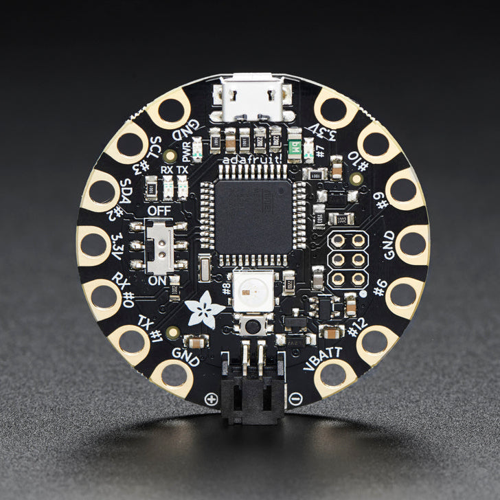 A circular electronic circuit board with a central microchip, various electronic components, and gold-plated circular pads around the edge, photographed on a dark gray background.