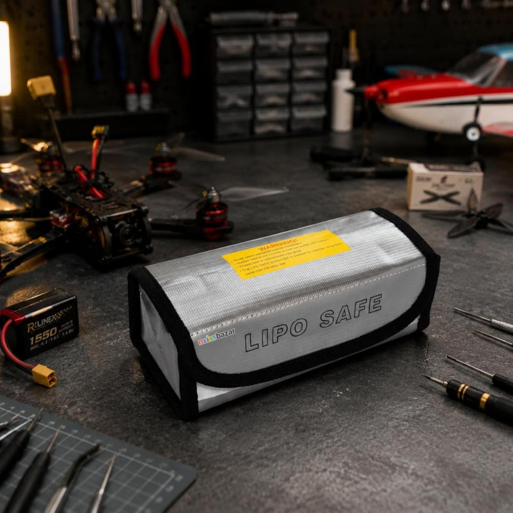 A silver and black Lipo Safe battery case with a yellow warning label, resting on a dark workbench surrounded by RC drone parts and tools.