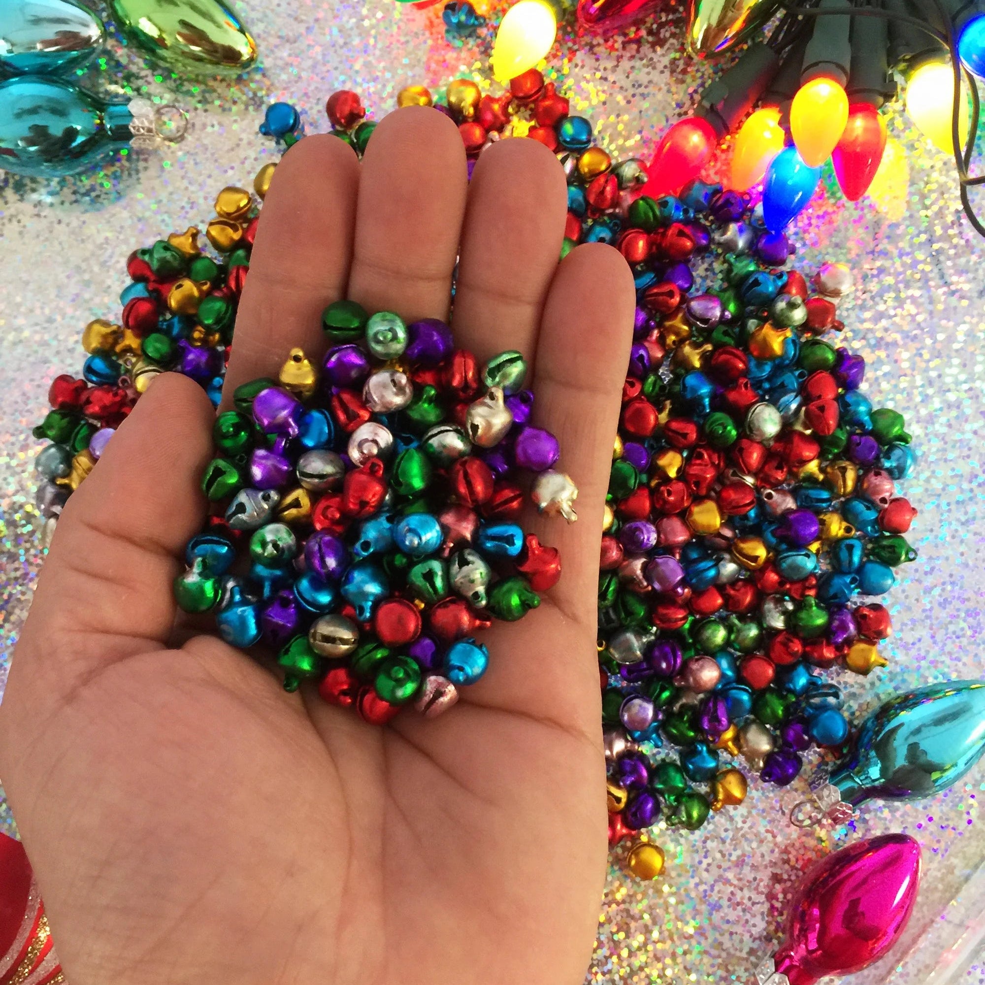 A hand holding a pile of small, shiny, metallic Christmas jingle bells in multiple colors, with more bells and colorful Christmas lights scattered in the background on a glittery surface.